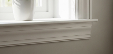 A white potted plant sits on a sunlit windowsill with ornate molding, casting soft shadows. The scene conveys a serene and minimalistic atmosphere.