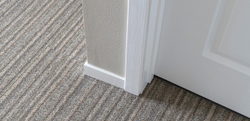 Close-up of a corner in a room with gray striped carpet meeting a beige wall. A white door partially opens, revealing a clean, modern interior.