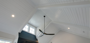A modern, white vaulted ceiling with exposed beams, featuring a large black ceiling fan. Natural light streams through upper windows, creating an airy feel.
