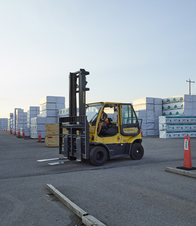 A person operates a yellow forklift on a paved lot, surrounded by large stacks of wrapped pallets. The scene is industrial and orderly, under a clear sky.