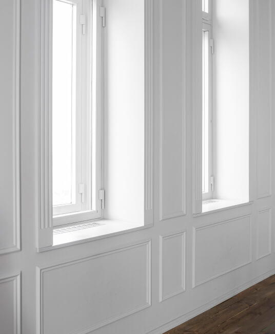 Bright, minimalist interior with tall, white-framed windows on a paneled wall, casting soft light onto a wooden floor, conveying serenity.