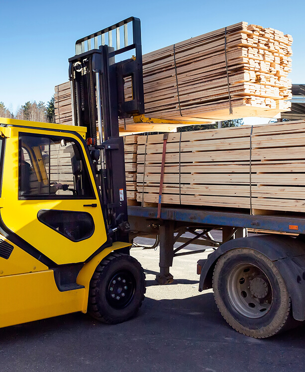A yellow forklift raises a stack of lumber onto a blue flatbed truck under a clear blue sky. The setting is industrial and efficient.