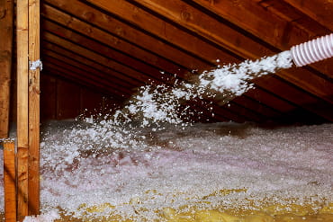 A hose sprays white insulation material into a wooden attic, covering the floor. The scene is focused on home improvement and energy efficiency.