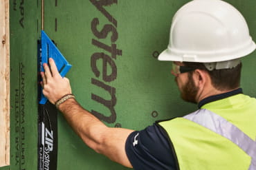 Worker applying ZIP System flashing tape with a hand roller to seal a seam on green sheathing panel.