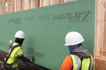 Two workers in hard hats installing ZIP System insulated R-sheathing panels on a wood-framed wall.