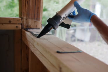 Close-up of gloved hand applying ZIP System liquid flash sealant to a wood corner joint with a caulking gun.