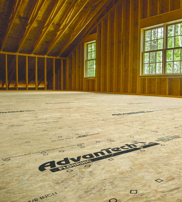 Unfinished attic room with AdvanTech subfloor panels installed over floor joists, exposed roof rafters, and two large windows letting in natural light.