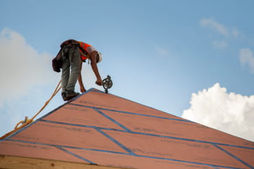 Worker on a steep roof installing ZIP System roof sheathing panels with taped seams, harnessed for safety.