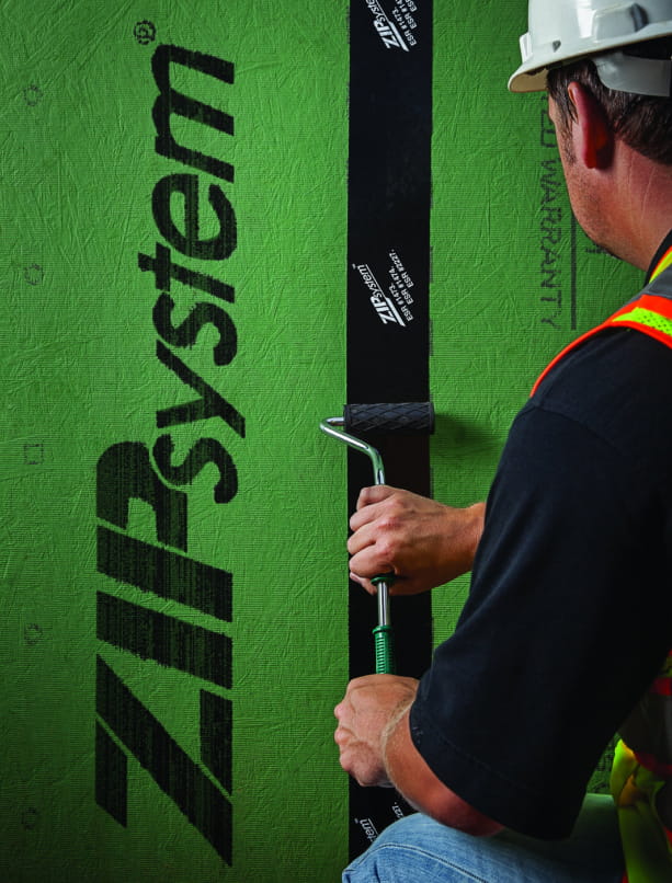 Worker in safety vest rolling ZIP System tape onto a seam of green sheathing panel with a J-roller.